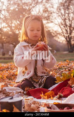 Das kleine Mädchen, 3-4 Jahre alt, sitzt draußen im Park und picknickt in den Strahlen der untergehenden Sonne und hält roten Apfel in den Händen. Dunkelhaarige Kind in Kleidung von Stockfoto