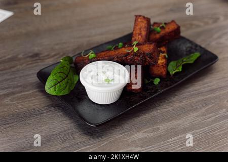 Leckere, würzige Schwarzbrotcroutons mit Knoblauchsauce, Salz und Basilikum auf schwarzem Teller auf Holztisch im Café. Junk Food Stockfoto