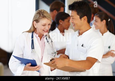 Sie lernen ziemlich schnell. Zwei Mediziner schauen sich ein Clipboard mit einer Reihe von Kollegen im Hintergrund an. Stockfoto