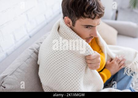 Blick aus der Ferne auf einen jungen Mann, der mit einer Decke bedeckt ist und auf dem Sofa sitzt und sich zu Hause warm werden kann, Stockbild Stockfoto