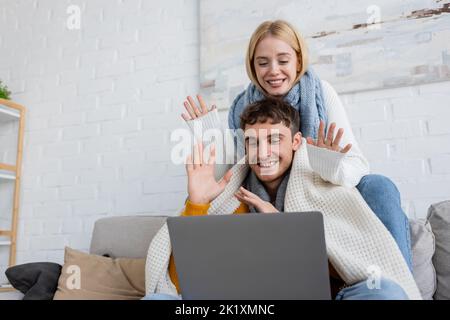Fröhliches junges Paar in Schals winkende Hände während Videoanruf auf Laptop, Bild Stock Stockfoto