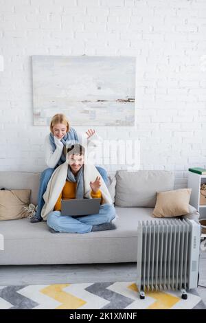 Fröhliches Paar mit Schals und Pullovern, die sich während eines Videoanrufs auf dem Laptop die Hände winken, Stockbild Stockfoto