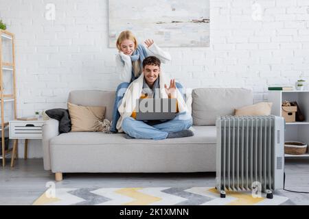 Glückliches junges Paar in Schals und Pullovern, die sich während eines Videoanrufs am Laptop die Hände winken, Bild im Stock Stockfoto