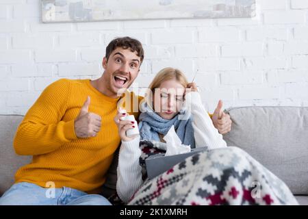 Fröhlicher Mann zeigt Daumen in der Nähe von kranken blonden Frau mit Tissue-Box auf dem Sofa sitzend, Stock Bild Stockfoto