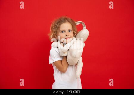 Kleines, wenig expressives, positiv lockiges blondes Mädchen, das flauschiges weißes Kaninchen in den Händen hält, betrachten Sie die Kamera auf der roten Ansicht Stockfoto