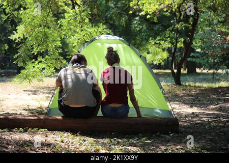Ein junges Paar, das auf einem Wald vor einem grünen Zelt in einem von hinten gefangenen Wald sitzt Stockfoto