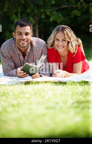 Ein glückliches junges Paar, das ein Buch liest, während es auf dem Rasen in einem Park liegt. Stockfoto