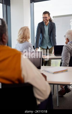 Ein fröhlicher Hipster-Pädagoge lehrt eine multikulturelle Gruppe älterer Studenten, wie man ein Projekt im Klassenzimmer schreibt. Stockfoto