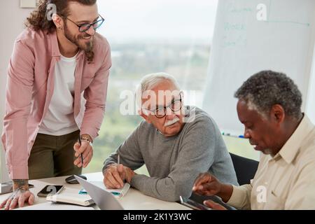 Ein junger Lehrer lehrt eine kleine Gruppe multikultureller älterer Schüler, wie man Technologien nutzt. Stockfoto