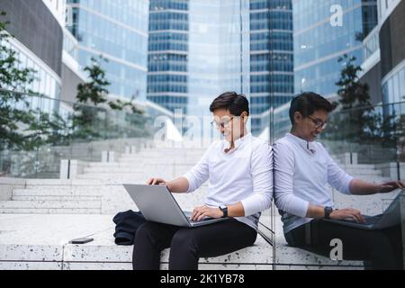 Junger Geschäftsmann, der im Freien in der Stadt sitzt und an einem Laptop arbeitet Stockfoto