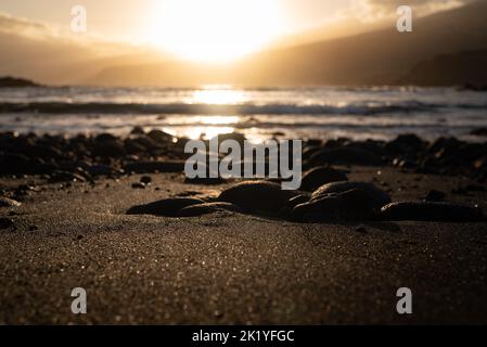 Eine meist verschwommene Nahaufnahme des schwarzen Sandstrandes mit vulkanischen Felsen bei Sonnenuntergang Stockfoto