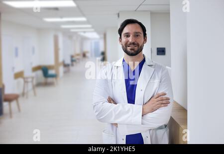 Porträt eines glücklichen männlichen Arztes, der mit gekreuzten Armen im Krankenhausflur steht. Gesundheitsfachkraft Stockfoto