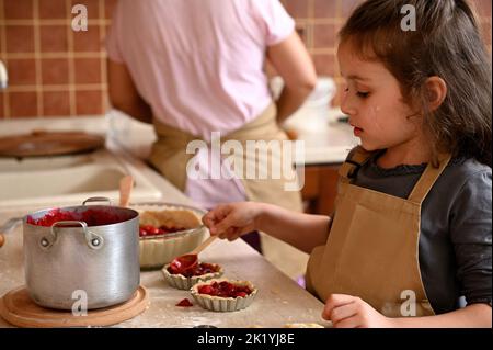 Kleines Mädchen in Kochschürze füllt Formen mit gerolltem Teig und Kirschen, bereitet Tartlets nach Familienrezept Stockfoto