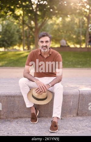 Ein entspannter Mann mittleren Alters, der einen Hut hält und auf die Kamera schaut, während er auf einer Bank in einem Park sitzt. Stockfoto