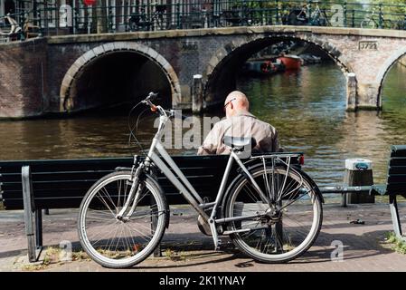 Amsterdam, Niederlande - 7. Mai 2022: Rückansicht eines unbekannten alten Mannes, der mit dem Fahrrad am Ufer des Kanals sitzt Stockfoto