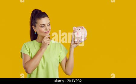 Junge Frau, die Zweifel an der Geldanlage hat, hält Sparschwein und denkt nach Stockfoto