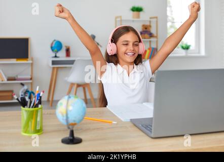 Mädchen hebt glücklich und siegreich ihre Hände, nachdem sie das Schulprojekt zu Hause auf einem Laptop abgeschlossen hat. Stockfoto