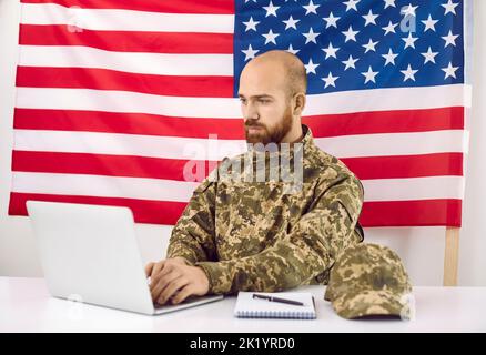 Amerikanischer Soldat, der am Laptop arbeitete, während er am Schreibtisch saß und die US-Flagge im Hintergrund hatte Stockfoto