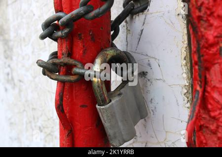 Ein altes Vorhängeschloss auf einer rostigen Kette, die um rote Rohre gewickelt ist. Stockfoto