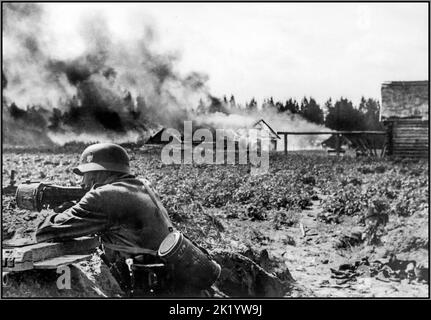 WW2 OPERATION BARBAROSSA Gegenparteiliche Aktion von Whermacht Nazi-deutschen Soldaten an der Ostfront. Im Vordergrund ein deutscher Soldat in Grabenstellung, der ein MG-34-Maschinengewehr mit einem Trommelmagazin abfeuerte. Ein brennendes Dorf im Hintergrund. 1941 Stockfoto