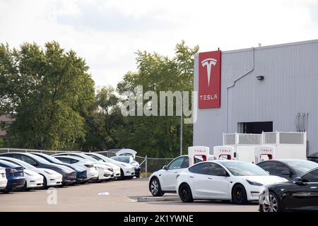 Das Tesla Logo befindet sich an der Seite eines Tesla Händlers und einer Ladestation für den Kompressor, die mit aufladenden Autos und neuen Fahrzeugen auf dem Parkplatz gefüllt ist. Stockfoto