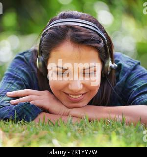Den Sommer wegwallen. Porträt einer attraktiven jungen Frau, die auf dem Gras liegt und Musik hört. Stockfoto