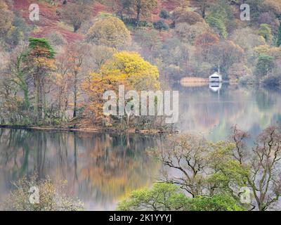 Herbstlaub spiegelt sich an einem ruhigen Novembermorgen im Wasser von Rydal wider, wo das berühmte Bootshaus die Szene verankert. Stockfoto