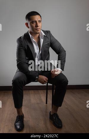 Junger latein mit kurzen Haaren und Bart, Business-Kleidung, Anzug, Hemd und Krawatte, Mode und Lifestyle im Studio auf einem Stuhl sitzend Stockfoto
