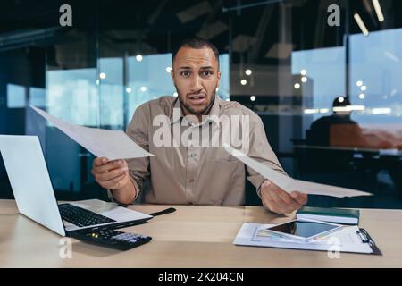 Porträt eines enttäuschten jungen hispanischen Finanziers, eines Geschäftsmannes, der verzweifelt die Kamera ansah und einen gescheiterten Finanzbericht über das Geschäft in Händen hielt, eines Mannes, der in Bürogebäude arbeitete. Stockfoto