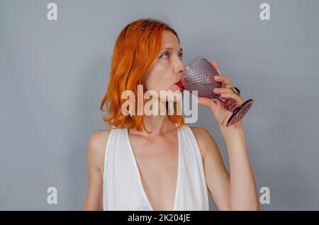 Porträt einer rothaarigen Frau in Weiß mit einem Glas Wein. Eine Frau trinkt aus Glas. Seitenansicht. Stockfoto