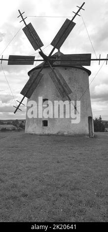 Schwarzweiß-Foto mit einer alten Windmühle, die auf einem Feld mit bewölktem Himmel sitzt Stockfoto