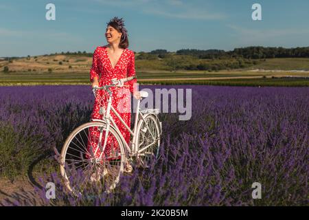 Eine junge Frau in einem roten Kleid steht mit einem Fahrrad in einem Lavendelfeld Stockfoto