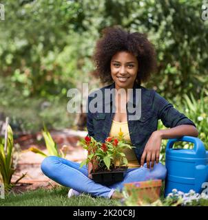 Immer ihren grünen Daumen schmutzig. Porträt einer glücklichen jungen Frau, die mit einem Tablett mit Pflanzen und Gartengeräten im Garten sitzt. Stockfoto