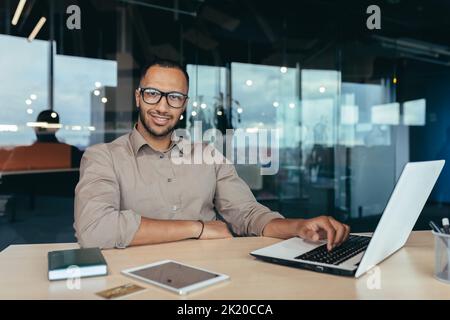 Porträt eines erfolgreichen Geschäftsmannes, eines Mannes mit Laptop, der im modernen Bürogebäude arbeitet, eines Startup-Unternehmers, der mit einem Laptop arbeitet, eine Brille trägt und lächelt und die Kamera anschaut. Stockfoto