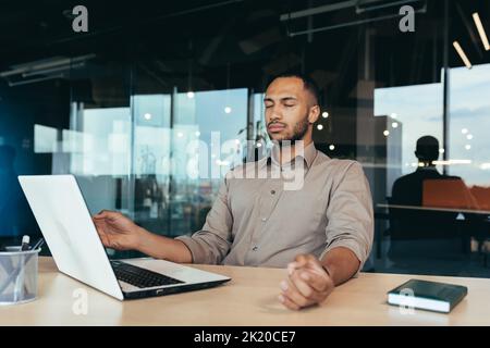 Erfolgreicher Geschäftsmann, der im modernen Bürogebäude arbeitet, meditiert mit geschlossenen Augen in Lotusposition auf einem Stuhl. Stockfoto