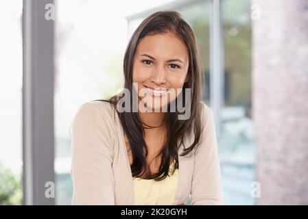 Girl-Next-Door-Charme. Eine glückliche junge Frau lächelt der Kamera zu, während sie zu Hause sitzt. Stockfoto