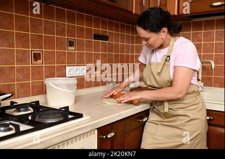 Schöne Frau in beigefarbener Kochschürze, die neben einer Küchenarbeitspüche steht und mit einem Nudelholz Teig für Brot ausrollt Stockfoto