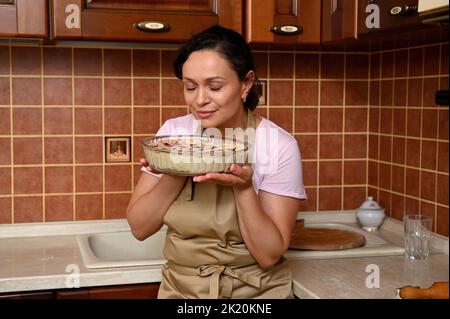 Eine wunderschöne dunkelhaarige Frau auf einer beigefarbenen Kochschürze hält einen rohen hausgemachten Kirschkuchen, bevor sie ihn in den Ofen legt Stockfoto