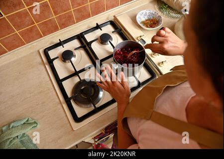 Blick von oben auf die hübsche Frau auf der Schürze des Küchenchefs, die Zucker mit Kirschen beträufelt, während sie karamellisierte Füllung für Kuchen zubereitet Stockfoto