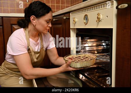 Eine wunderschöne Hausfrau mittleren Alters, die eine beige Kochschürze trägt und im Ofen in der heimischen Küche einen Kirschkuchen kocht Stockfoto