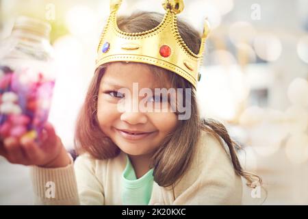 Möchten Sie ein paar Süßigkeiten der Königin? Ein süßes kleines Mädchen, das als Prinzessin verkleidet ist, während es zu Hause ein Glas Süßigkeiten hält. Stockfoto