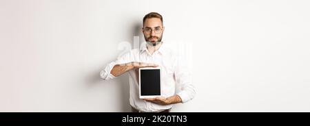 Selbstbewusster bärtiger Mann zeigt digitalen Tablet-Bildschirm, demonstriert App, stand über weißem Hintergrund Stockfoto