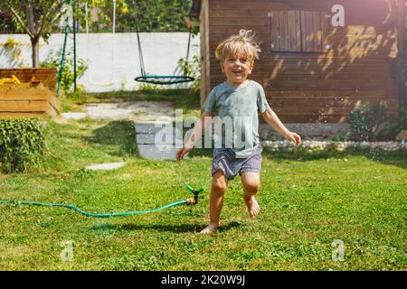 Spaß im Garten - hübscher Junge läuft um den Wasserregner herum Stockfoto