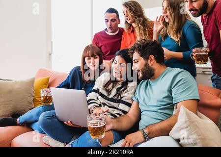 Eine Gruppe von glücklichen jungen Freunden, die Spaß haben, Inhalte von sozialen Netzwerken im Internet auf dem Laptop auf dem Sofa zu sehen und Bier zu trinken - Menschen, zusammen Stockfoto