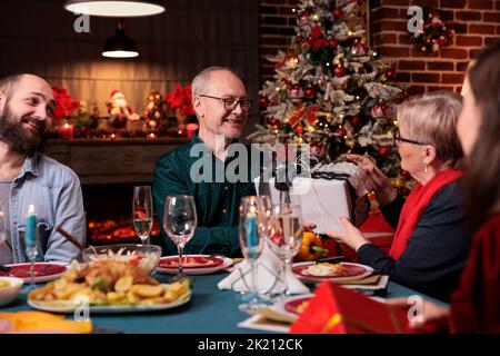 Familie feiert weihnachten, tauschen Geschenke an festlich servierten Tisch, essen traditionelle Gerichte zu Hause fest. Weihnachtsfeier mit Eltern, Menschen geben Geschenk-Boxen Stockfoto