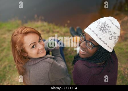 An einem Lieblingsort hängen und fangen. Hochwinkelporträt von zwei fröhlichen Teenagern, die neben einem See sitzen und heiße Getränke halten. Stockfoto