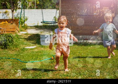 Spaß am heißen Sommertag zwei Kinder, Junge und Mädchen, laufen auf dem Rasen im Garten um den Wasserregner herum und hüpfen über Jets Stockfoto