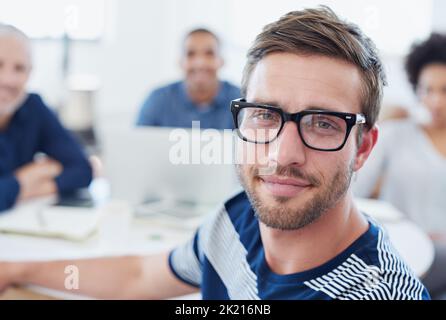 Hundert Prozent konzentrierten sich auf meine Karriere. Porträt eines jungen Büromitarbeiters, der an einem Tisch mit Kollegen im Hintergrund sitzt. Stockfoto