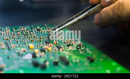 Grüne Leiterplatte mit allen elektrischen Komponenten, die von einem kaukasischen Ingenieur mit einer speziellen Pinzette repariert werden. Nahaufnahme, Handmontage. Hochwertige Fotos Stockfoto