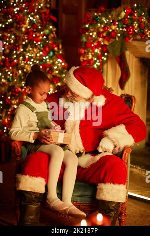 Vertikales Porträt von niedlichen afroamerikanischen Mädchen sitzen in Santas Schoß und halten Weihnachtsgeschenk in gemütlicher Umgebung Stockfoto